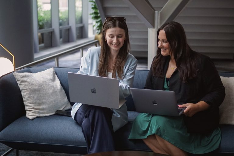 Two women are sitting in a corporate setting, sharing their computer screens with each other.