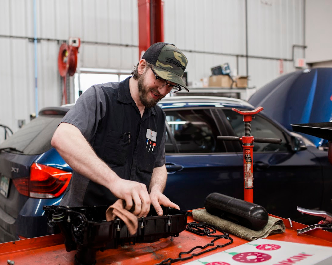A man is working on a car, wiping a car part with a rag. He is in a mechanic shop and is smiling while looking at what he is doing.